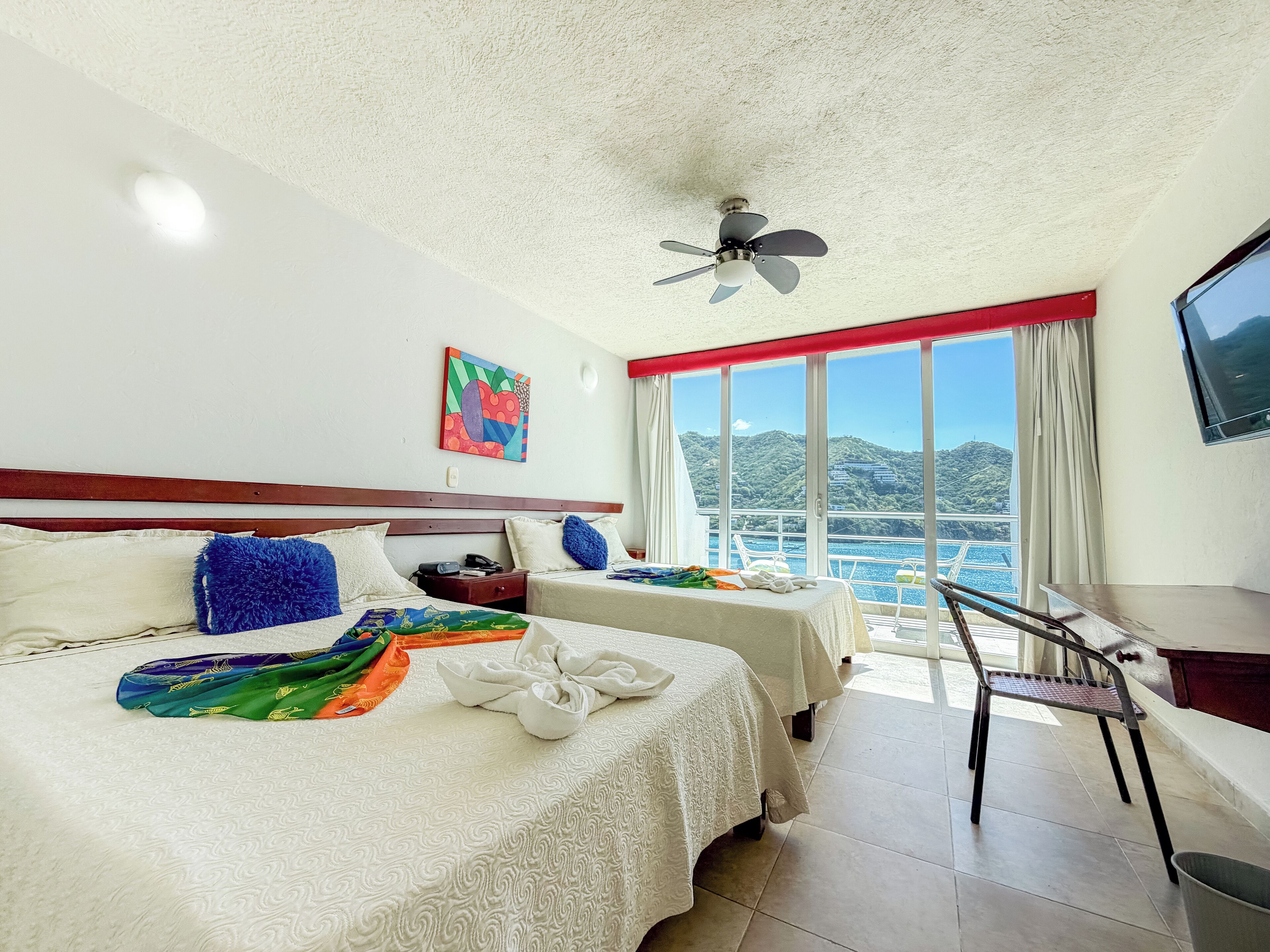 Ein Hotelzimmer mit zwei Betten, einem Schreibtisch, einem Stuhl, einem Deckenventilator, einem Gemälde und Meerblick.