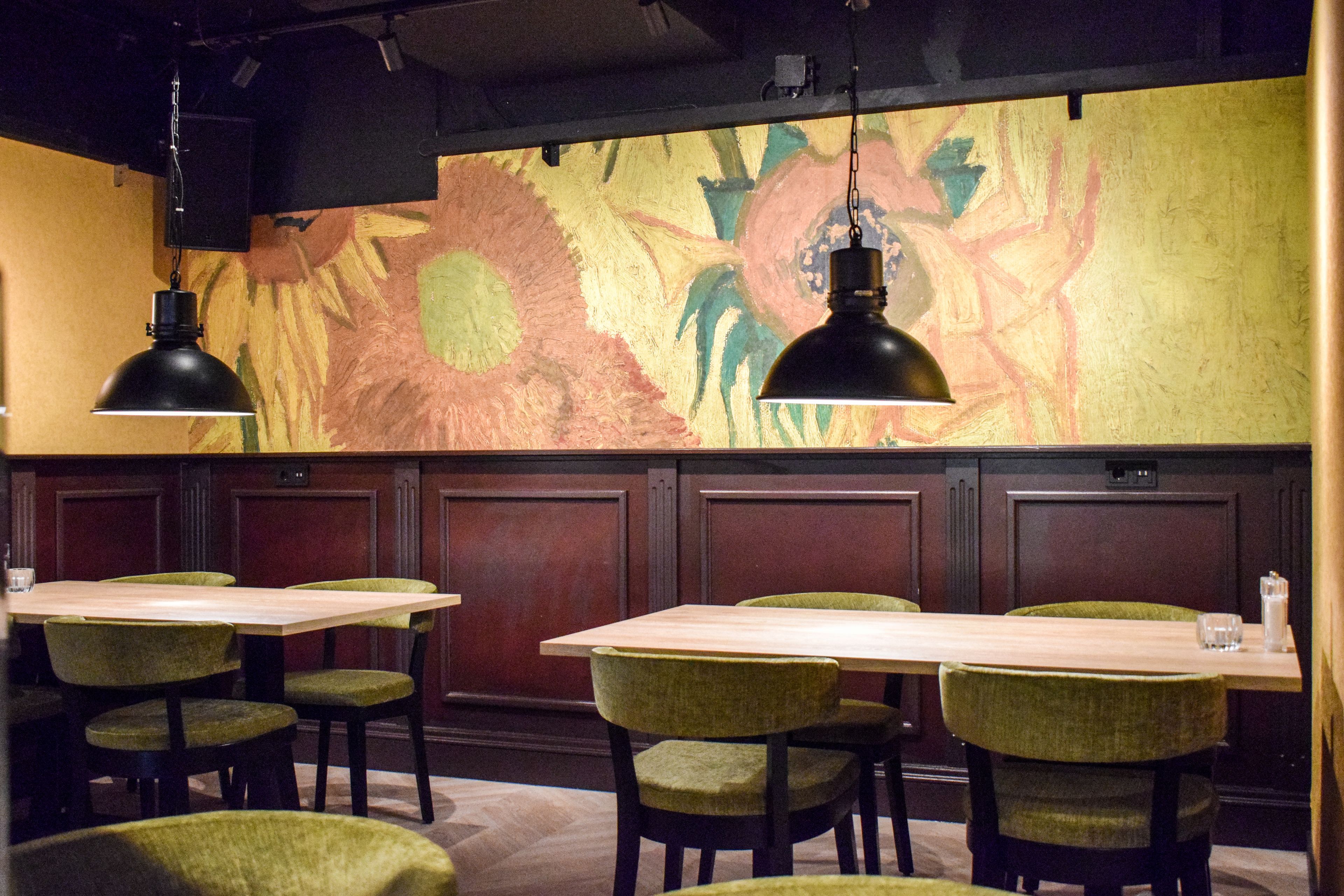 A restaurant interior with wooden tables and green upholstered chairs, a colorful abstract mural on the wall, and pendant lighting.
