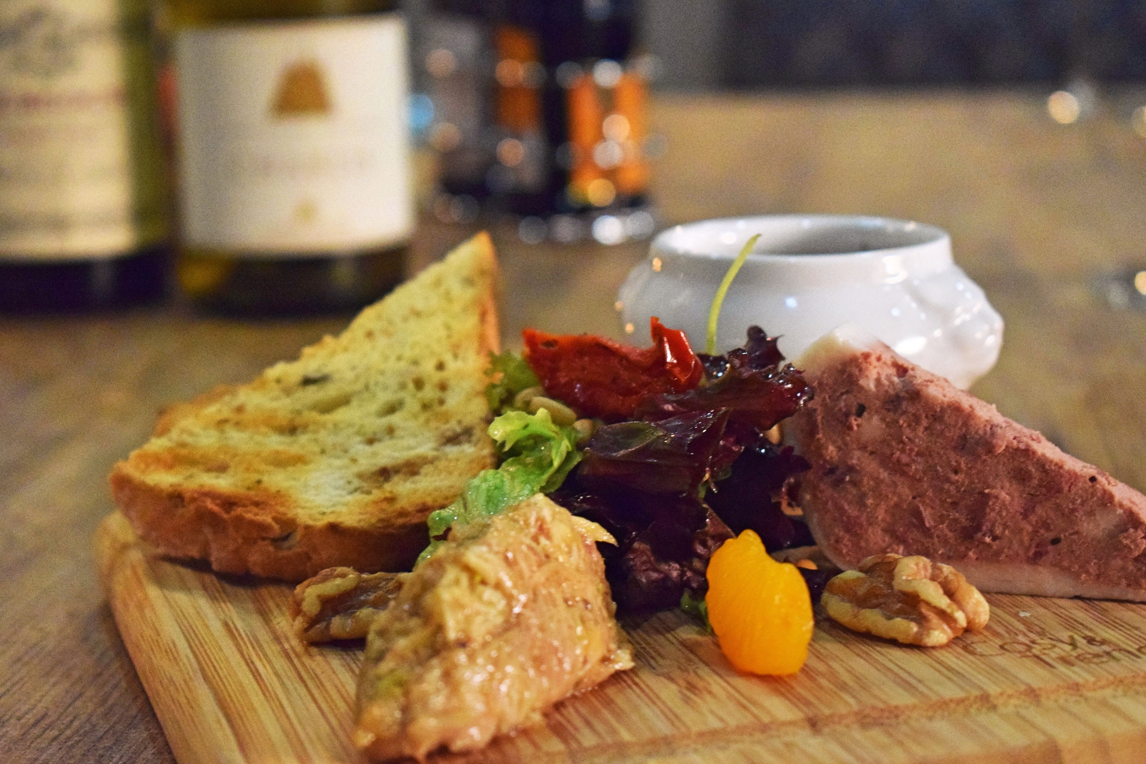 A wooden cutting board with various food items including bread, meat, nuts, and a small yellow fruit.