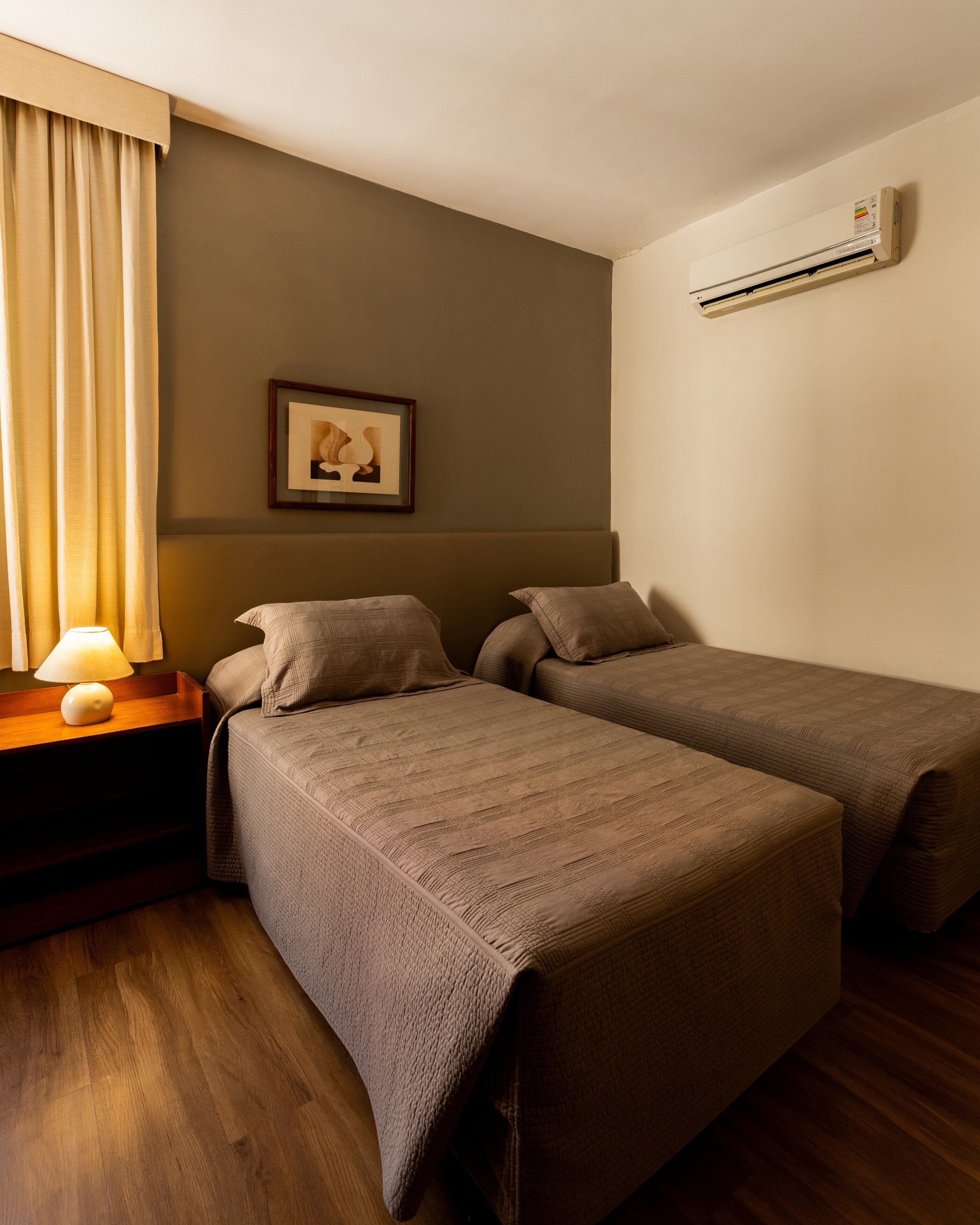 A hotel room with two beds, a nightstand with a lamp, a framed picture on the wall, and an air conditioning unit.
