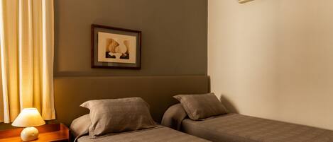 A hotel room with two beds, a nightstand with a lamp, a framed picture on the wall, and an air conditioning unit.
