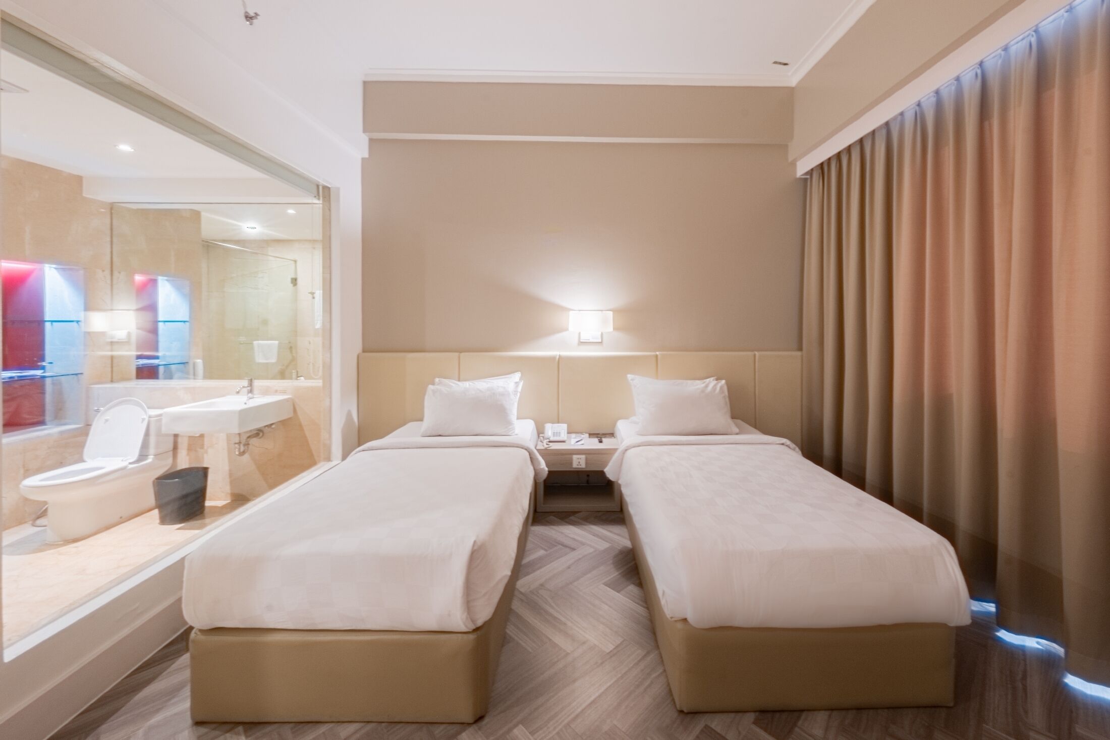 A hotel room with two beds, a bathroom with a mirror, sink, and toilet, and a large window with curtains.