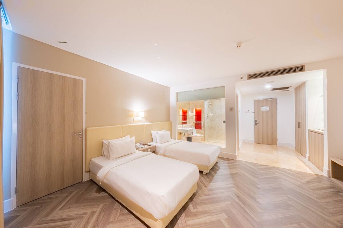Habitació d'hotel amb dos llits, un mirall gran i un bany visible a través d'una porta de vidre.
