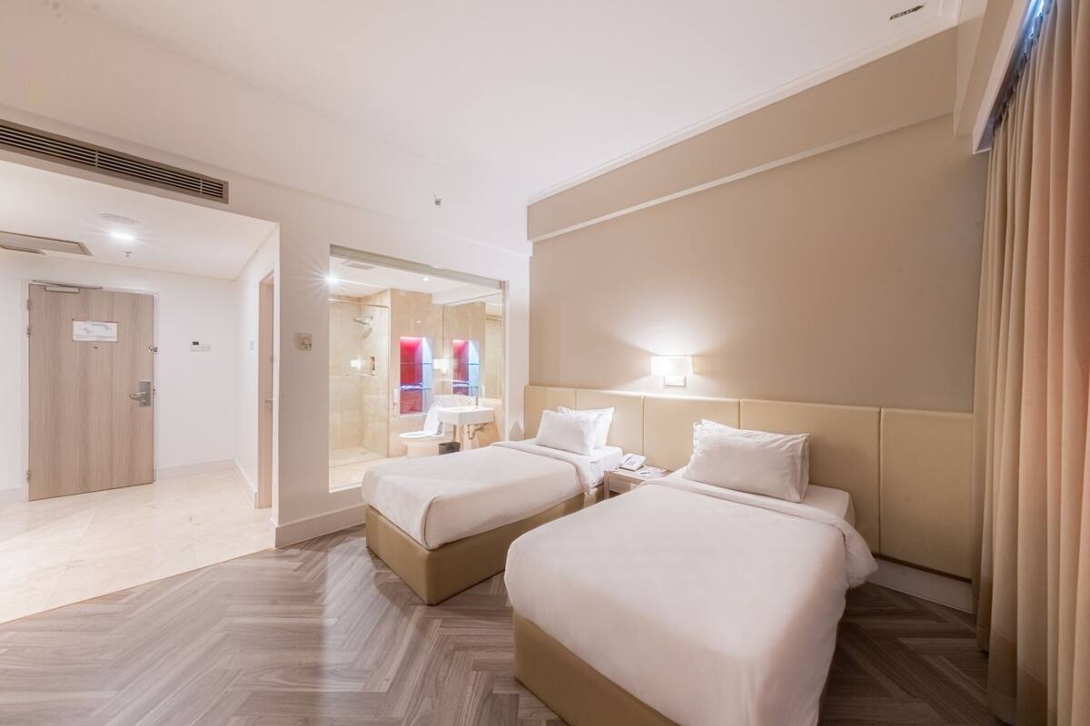 Una habitació d'hotel amb dos llits, un bany amb mirall i una porta que condueix a una altra habitació.