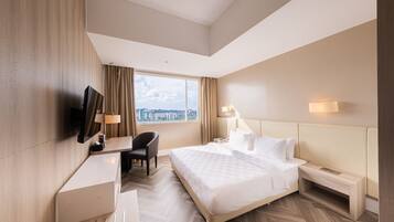 A modern hotel room with a large bed, a desk with a chair, a TV, and a window with a city view.