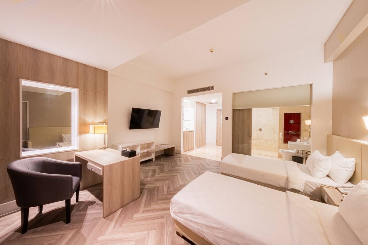 Una habitació d'hotel moderna amb un terra de fusta, un escriptori amb una cadira, un llit, un televisor fixat a la paret i un mirall gran.