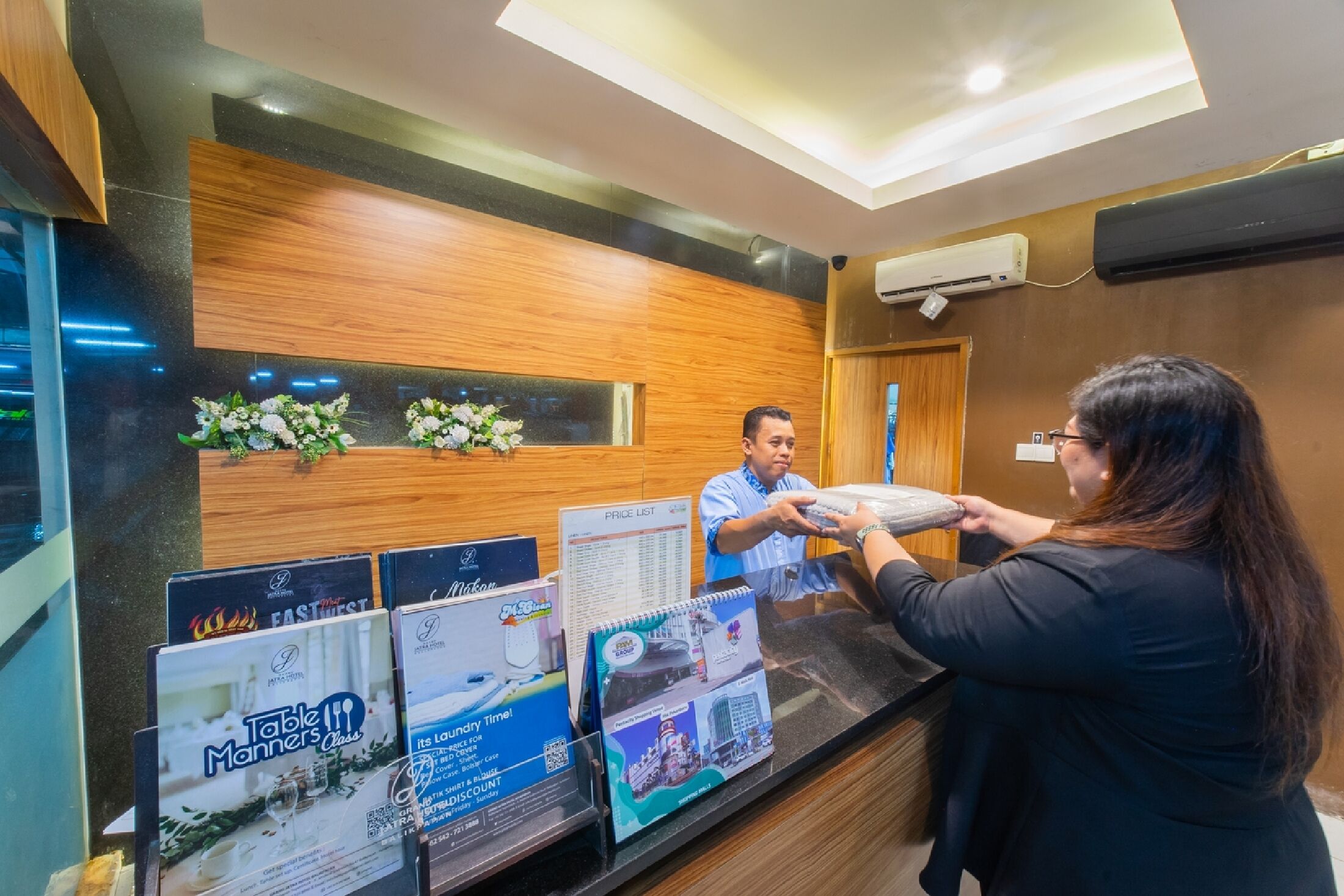 A reception desk with a man handing over a tablet to a woman. There are posters on the wall advertising table manners and a laundry service.