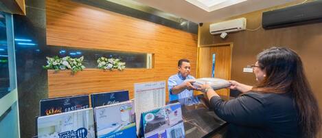 A reception desk with a man handing over a tablet to a woman. There are posters on the wall advertising table manners and a laundry service.