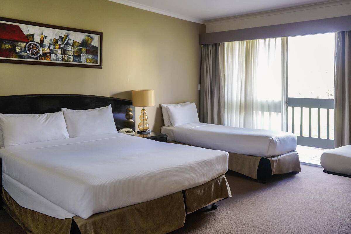 A hotel room with two beds, a painting on the wall, a lamp, and a view of the outdoors.