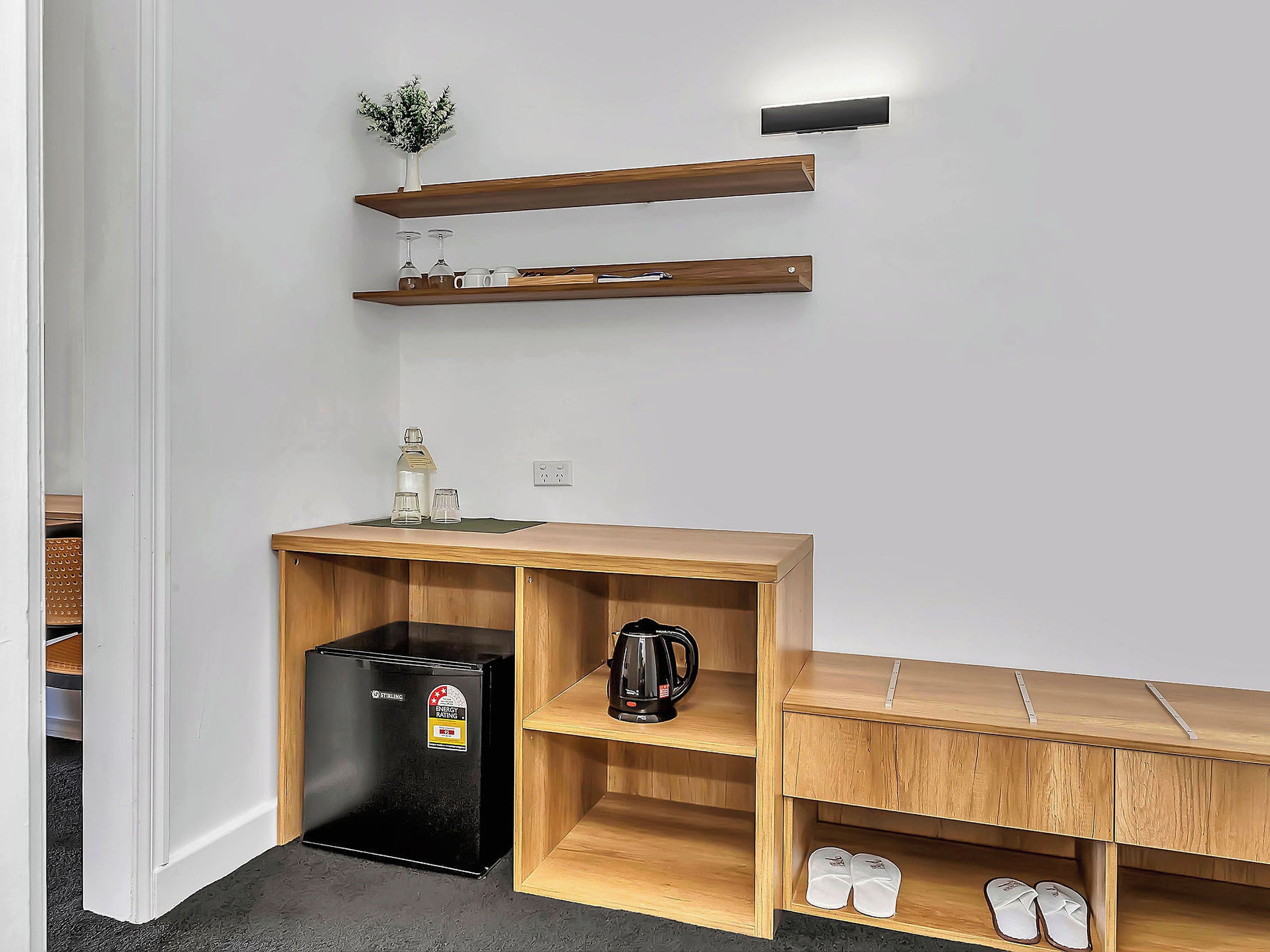 A compact kitchenette with a black refrigerator, wooden shelves, a bench, and a small plant.