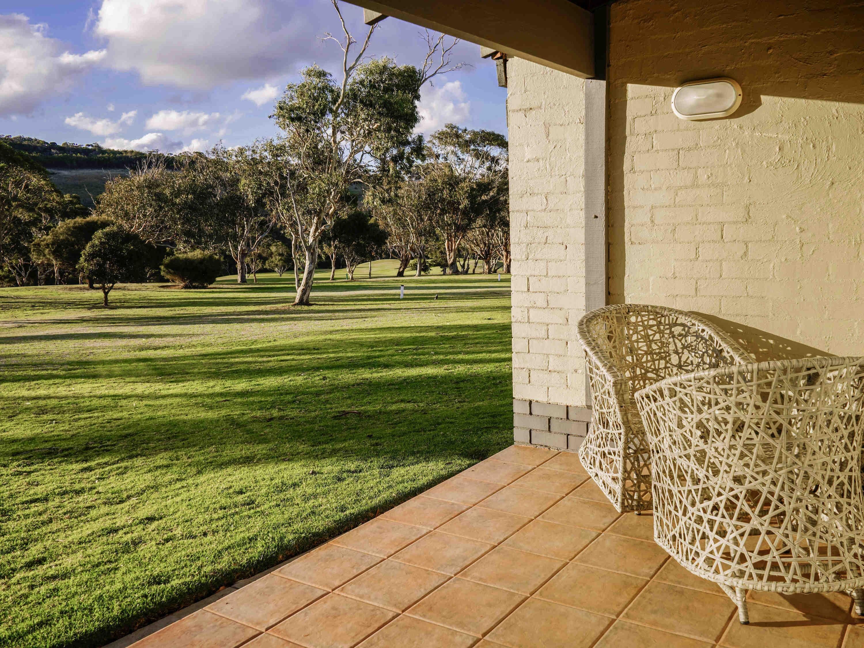 A patio area with a view of a grassy field and trees, a wicker chair, and a tiled floor.