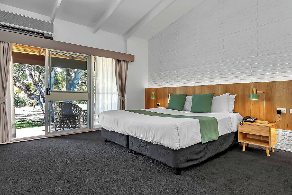 A hotel room with a large bed, a wooden headboard, a nightstand with a lamp, and a view through a glass door.