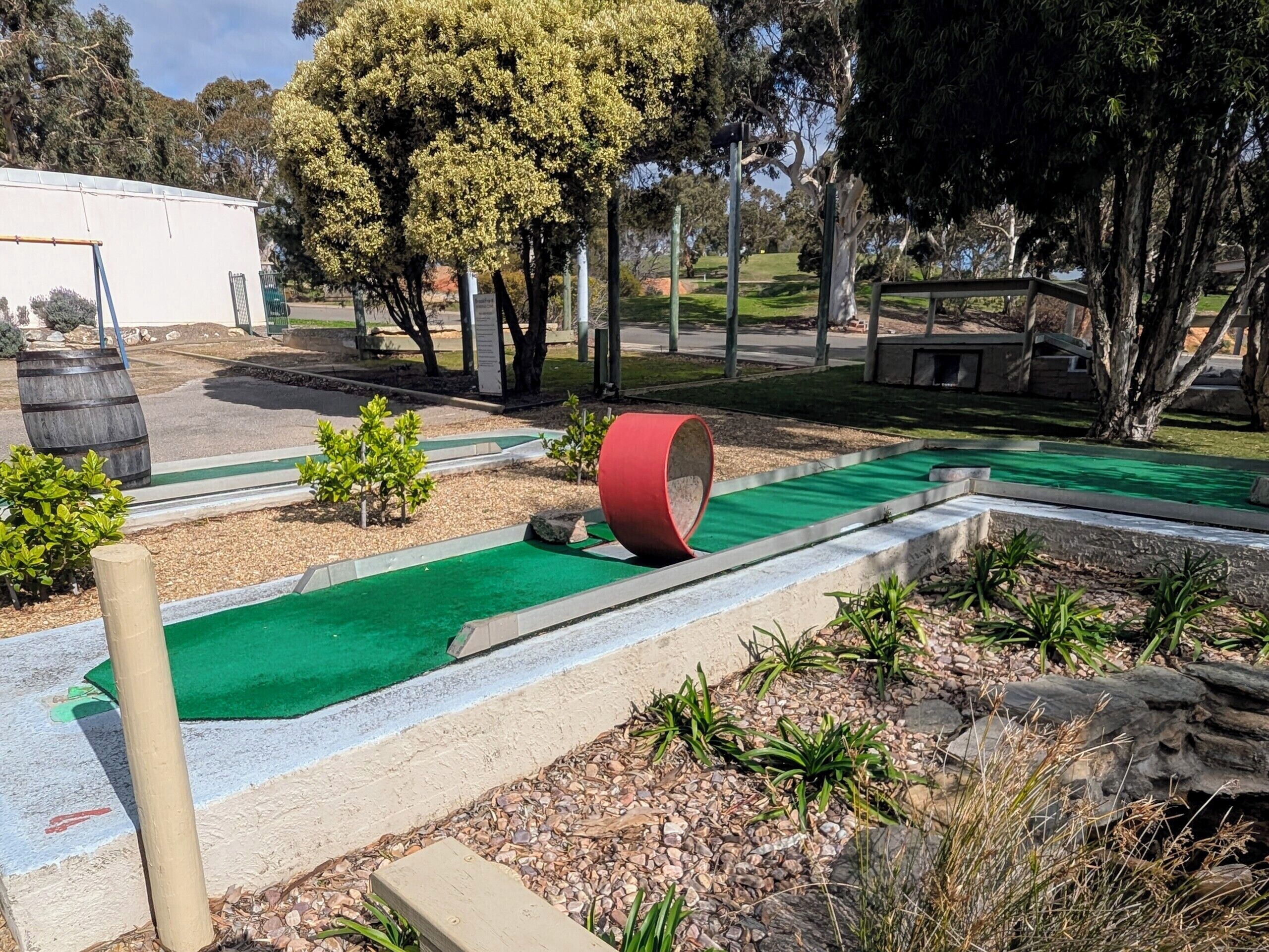 A miniature golf course with a green putting surface, a red cylindrical obstacle, and a small wooden bench.