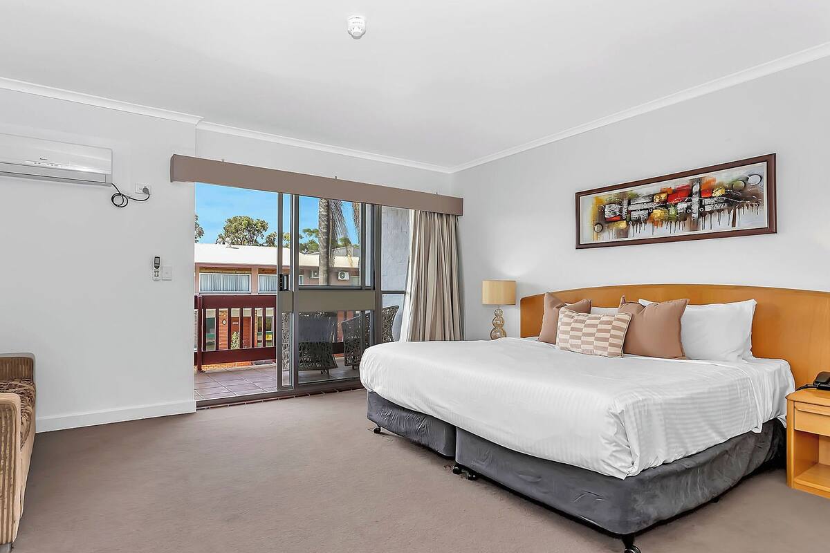 A hotel room with a large bed, a sliding glass door to a balcony, a painting on the wall, and a telephone on the nightstand.
