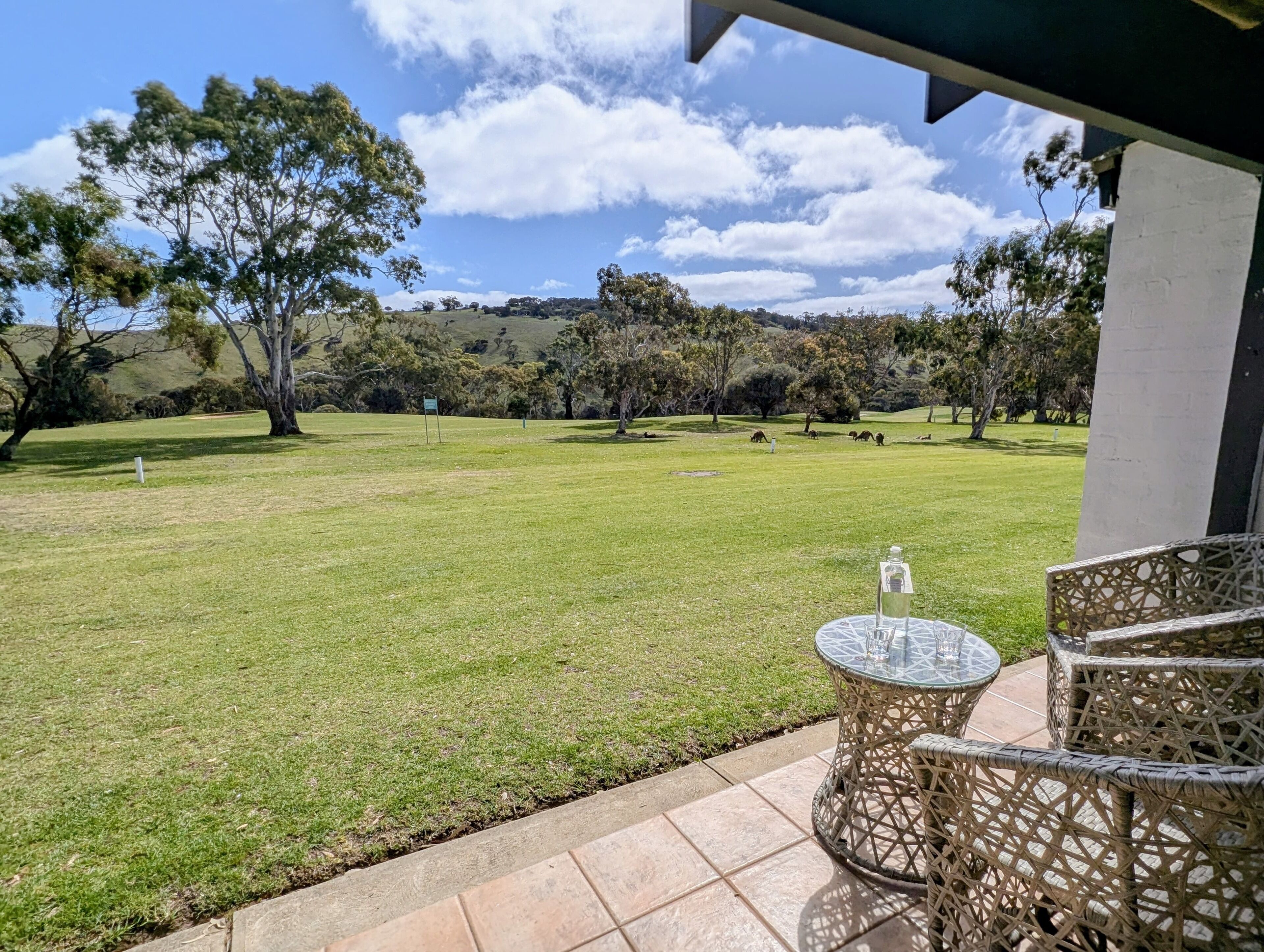 Un patio con muebles de mimbre y una mesa de vidrio, con vistas a un campo cubierto de césped, árboles y colinas al fondo.