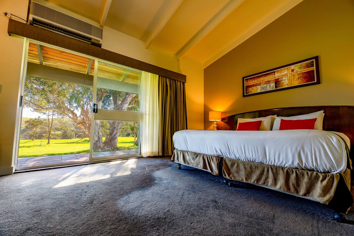 A hotel room with a large bed, a sliding glass door to a garden, and a painting on the wall.