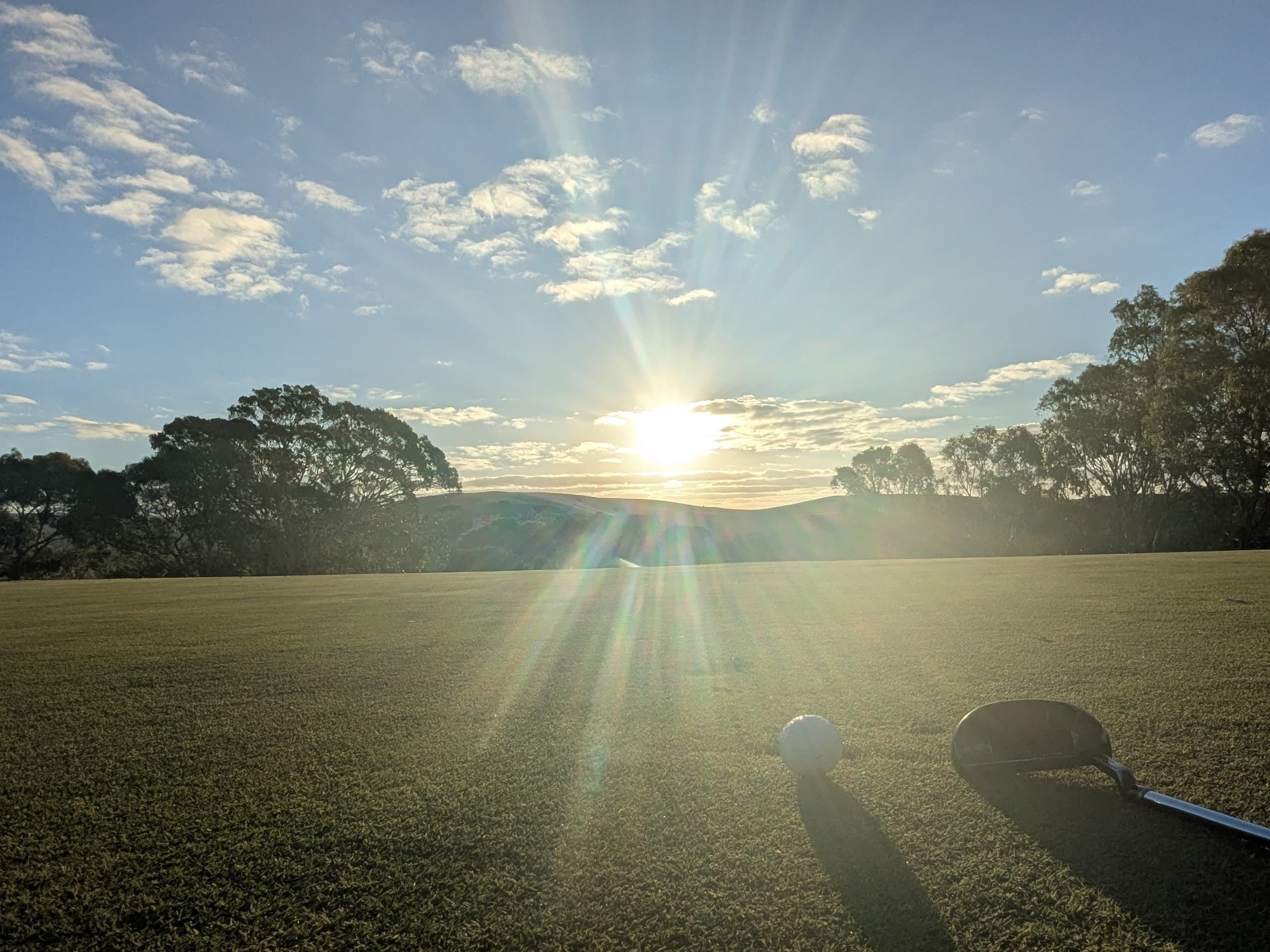 En golfbane med klar himmel, solstråler og en golfbold og kølle på græsset.