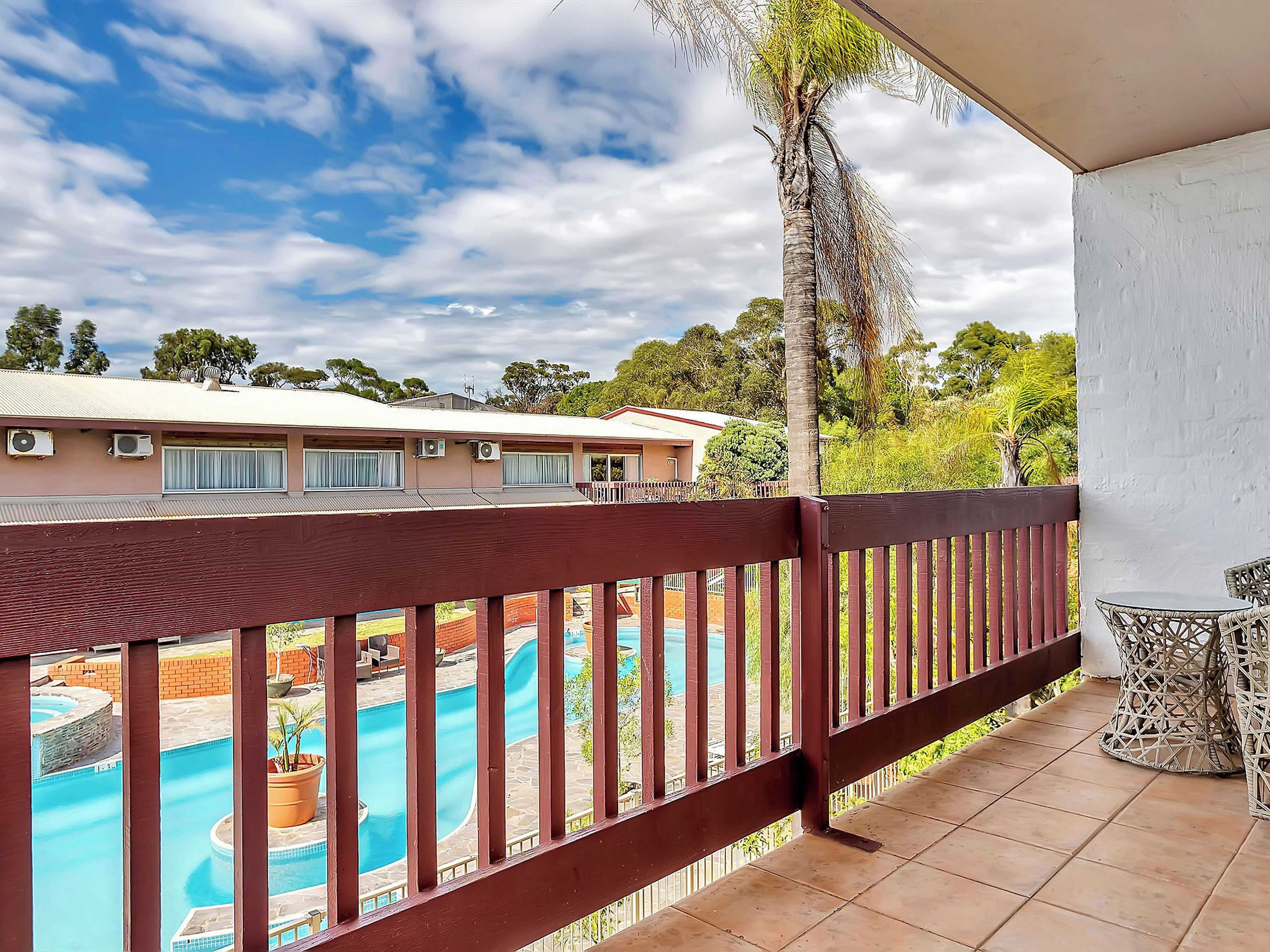 A balcony with a view of a swimming pool, a building with air conditioning units, and a palm tree.