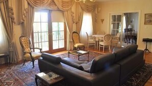 A living room with a black sofa, a wooden coffee table, and a patterned rug. There is a dining area with a table and chairs, and a chandelier hanging from the ceiling. - The Residence at Barossa Chateau (Lyndoch)