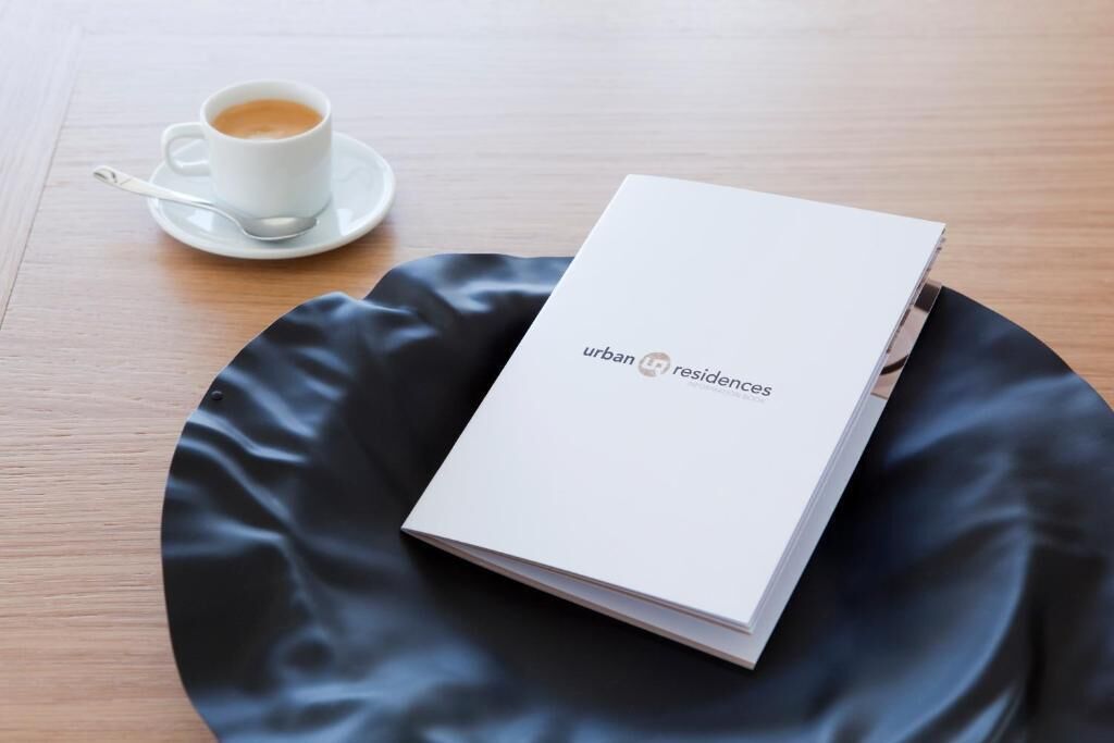 Um livreto branco com o título "urban residences" sobre uma superfície preta, ao lado de uma xícara de café branca com uma colher.
