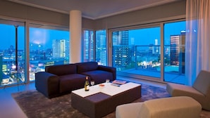 Una sala de estar moderna con un amplio ventanal que ofrece vista a la ciudad, un sofá de color oscuro, una mesa de centro blanca con una botella y vasos, y un sillón de tonos claros.