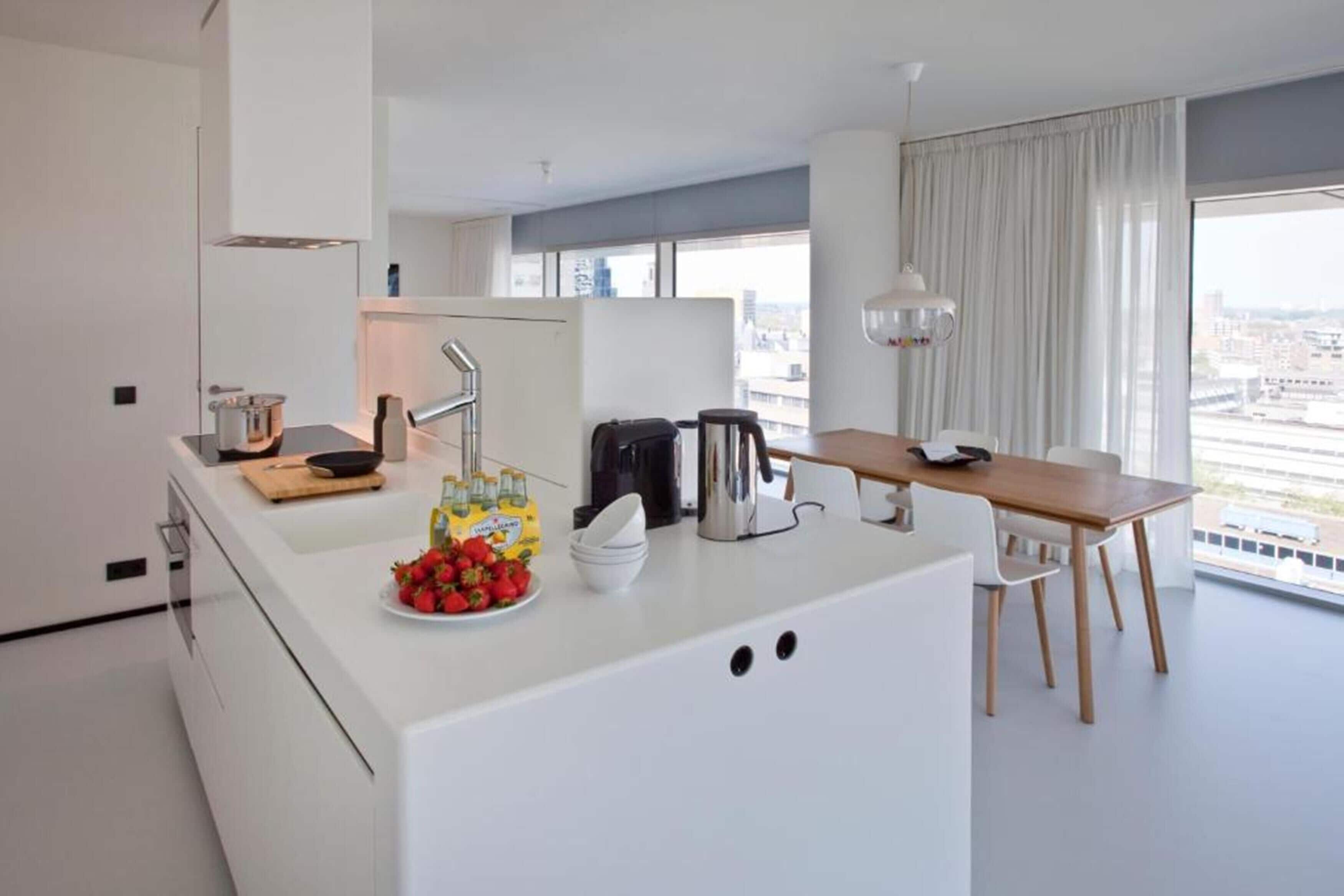 A modern kitchen with a central island, a dining area with a wooden table and chairs, and large windows offering a city view.