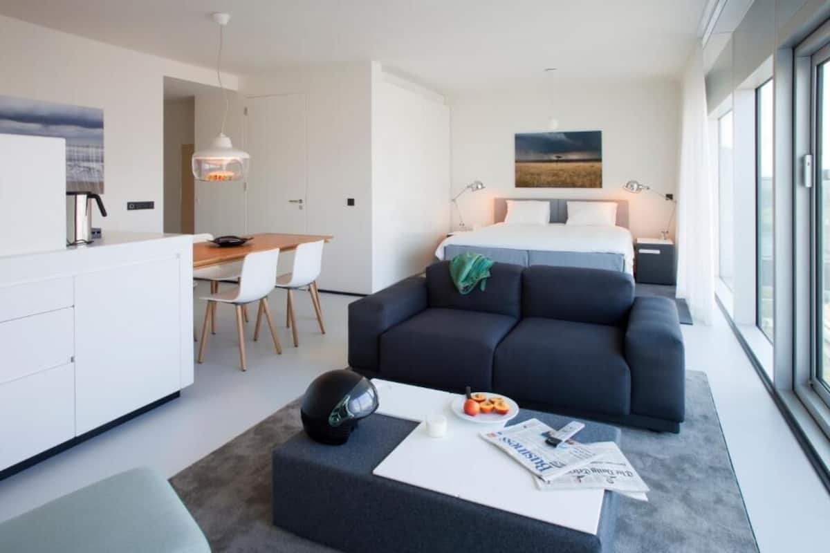 A modern hotel room with a bed, a sofa, a dining area, and a kitchen.