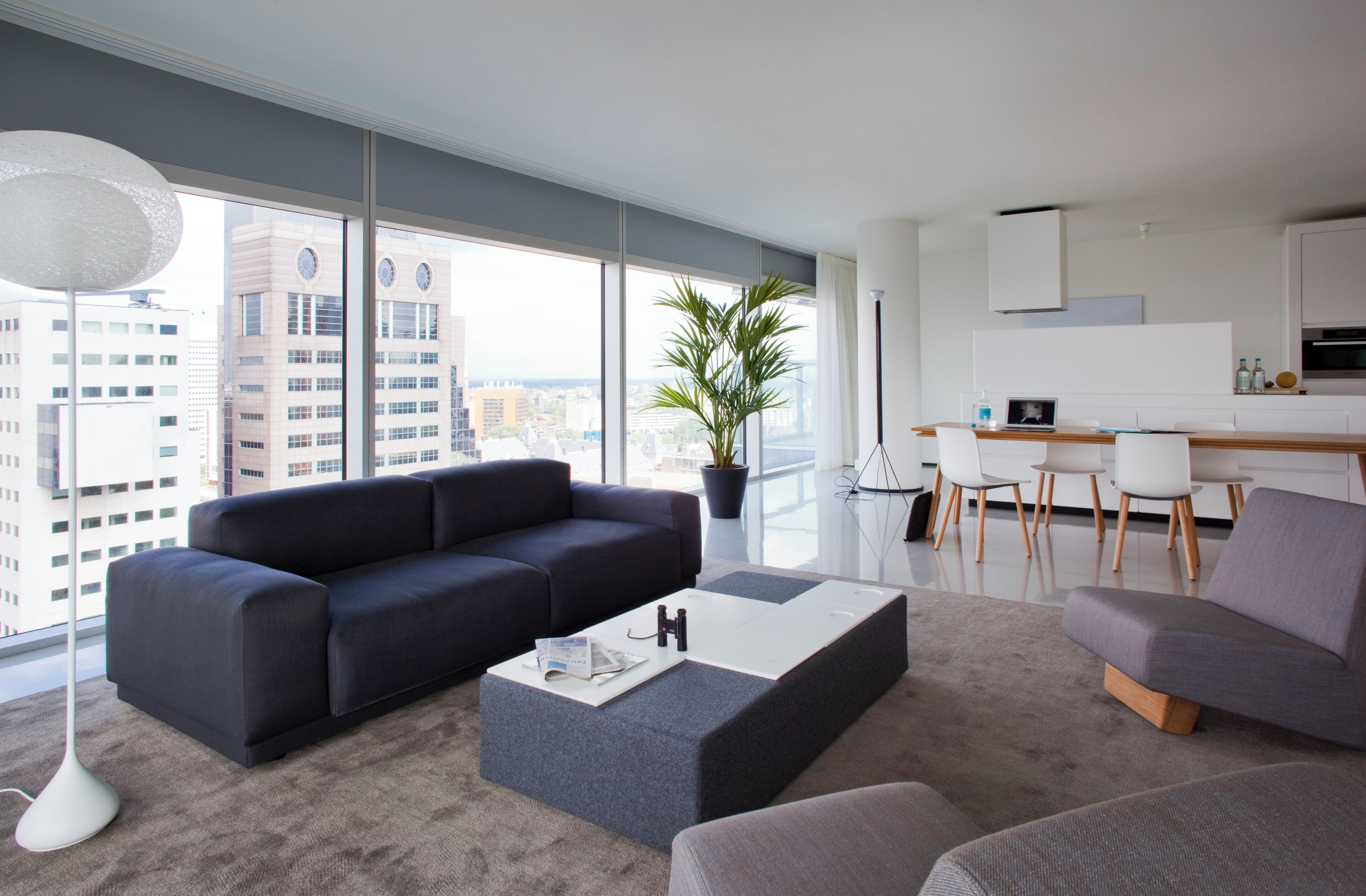 A modern living room with a large window, a black sofa, a grey ottoman, and a dining area with white chairs and a kitchen in the background.