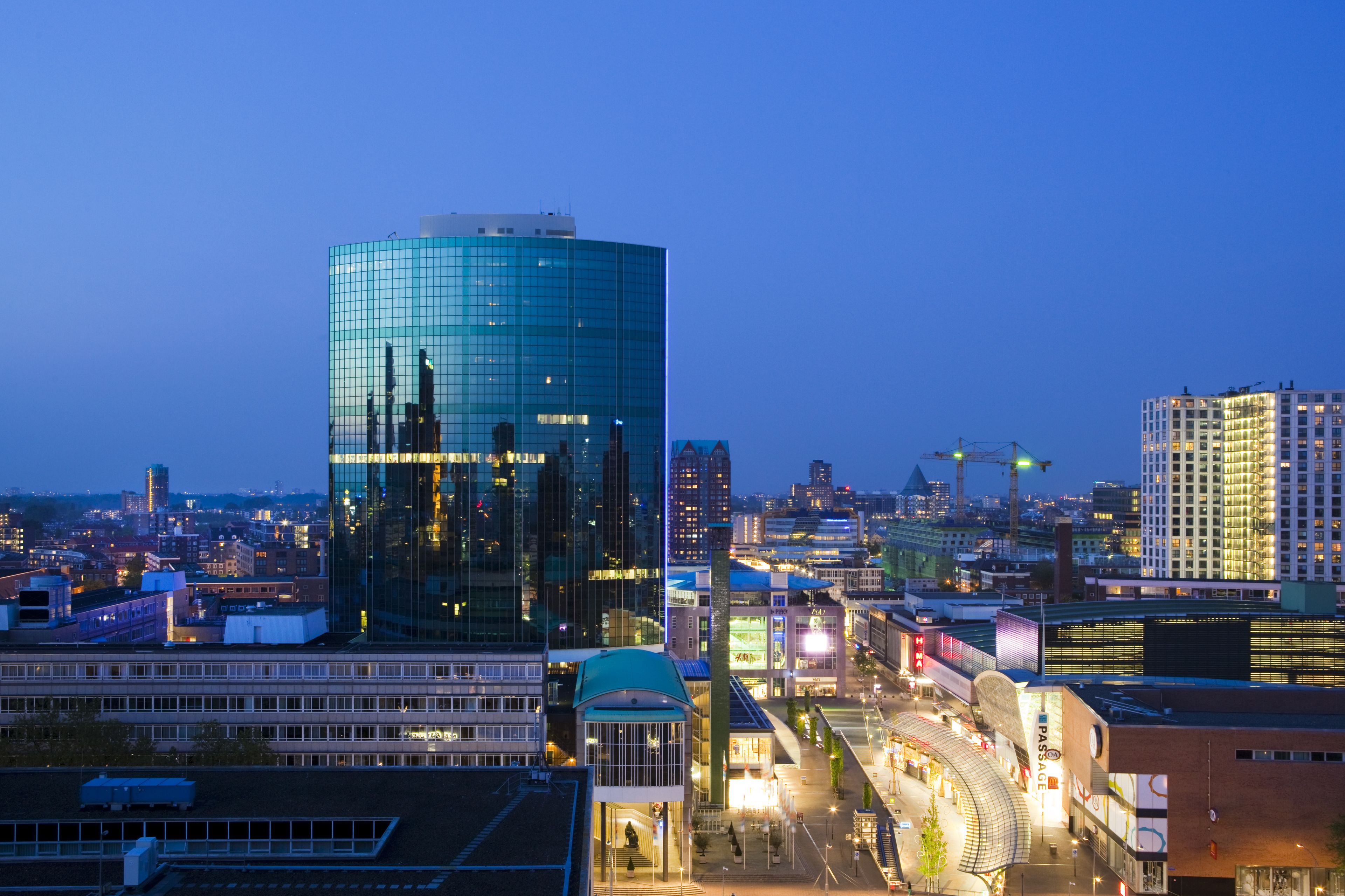 A cityscape at dusk with modern buildings, including a prominent glass skyscraper, and a well-lit commercial area with a curved building.