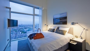 Habitación de hotel moderna con una cama grande, televisión, vistas a la ciudad y una mesita de noche con lámpara.