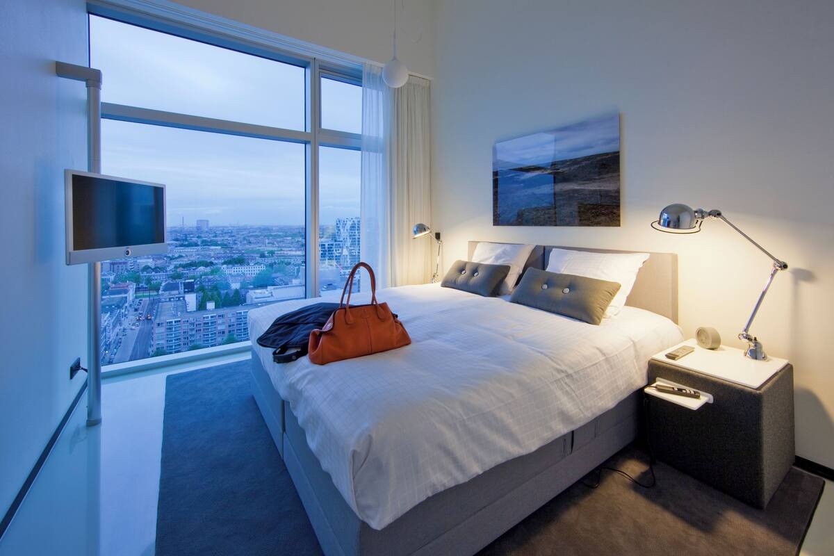 A modern hotel room with a large bed, a TV, a city view, and a nightstand with a lamp.