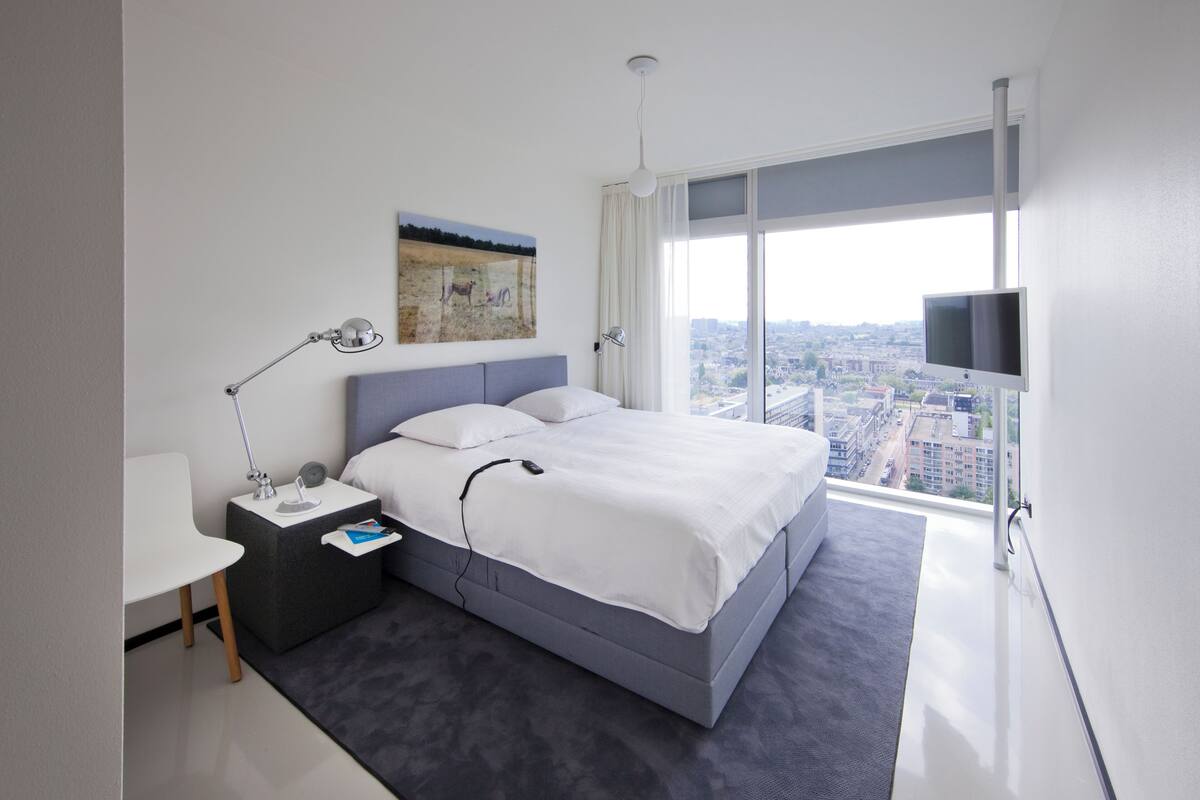 A modern hotel room with a large bed, a bedside table with a lamp, a flat-screen TV, and a city view.