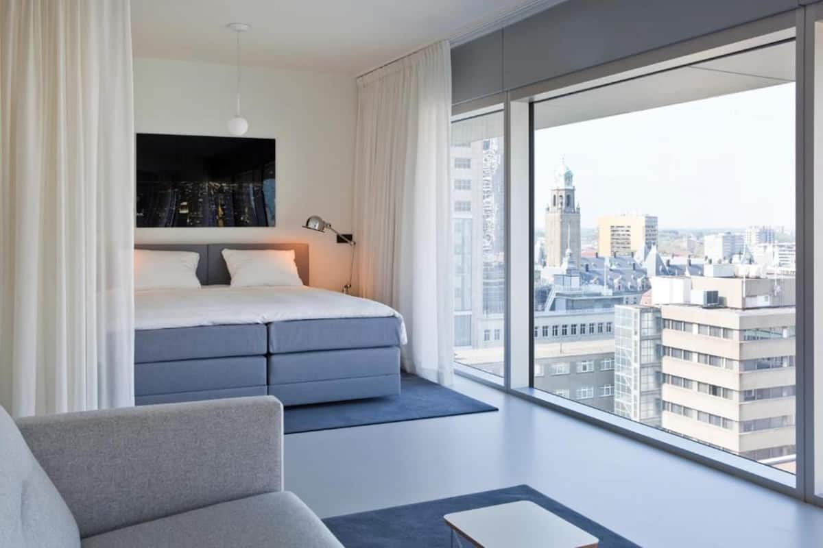 A modern bedroom with a large bed, a sofa, and a city view through floor-to-ceiling windows.