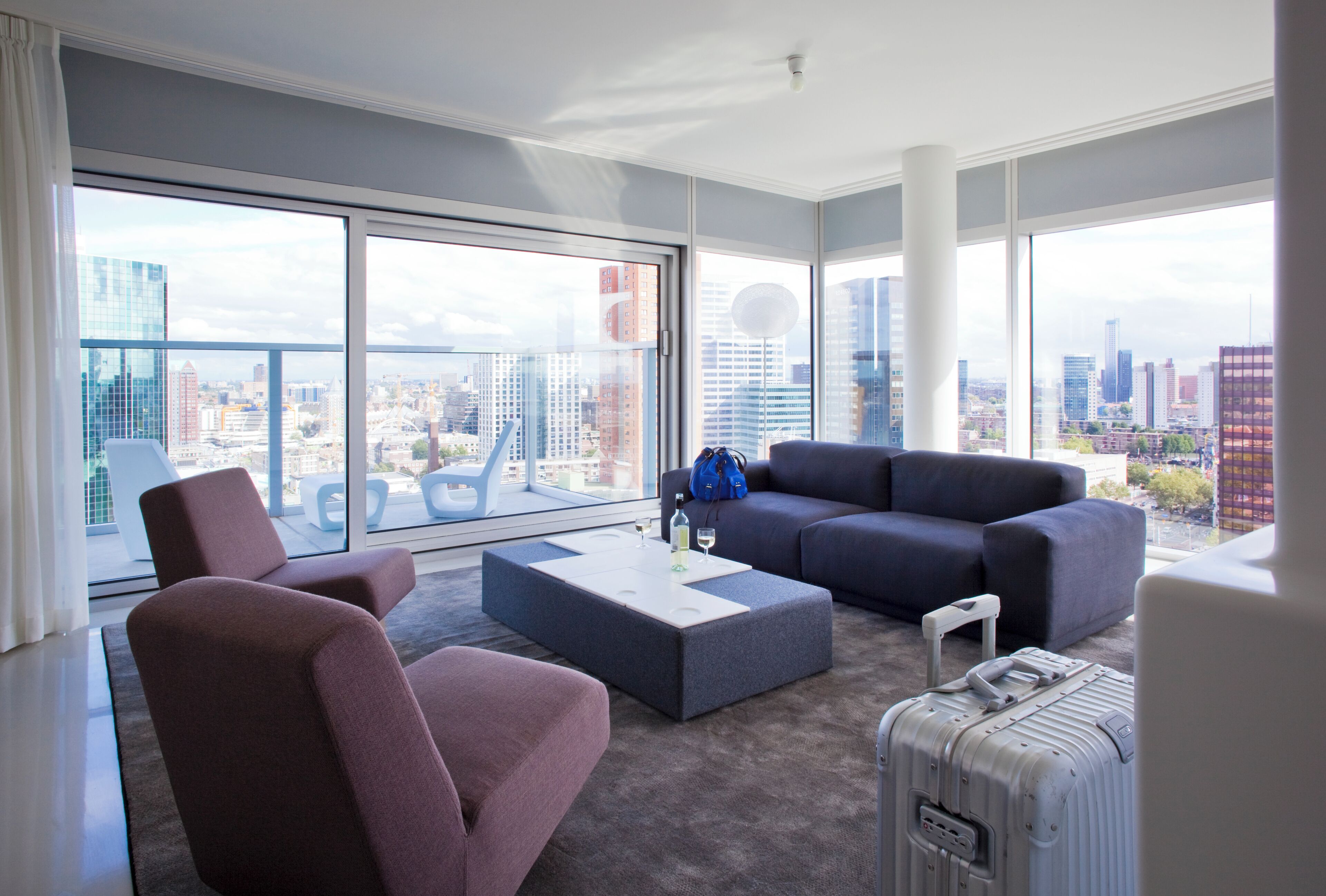Quarto de hotel moderno com uma grande janela que oferece vista da cidade, um sofá cinza, uma mesa pequena e uma mala.