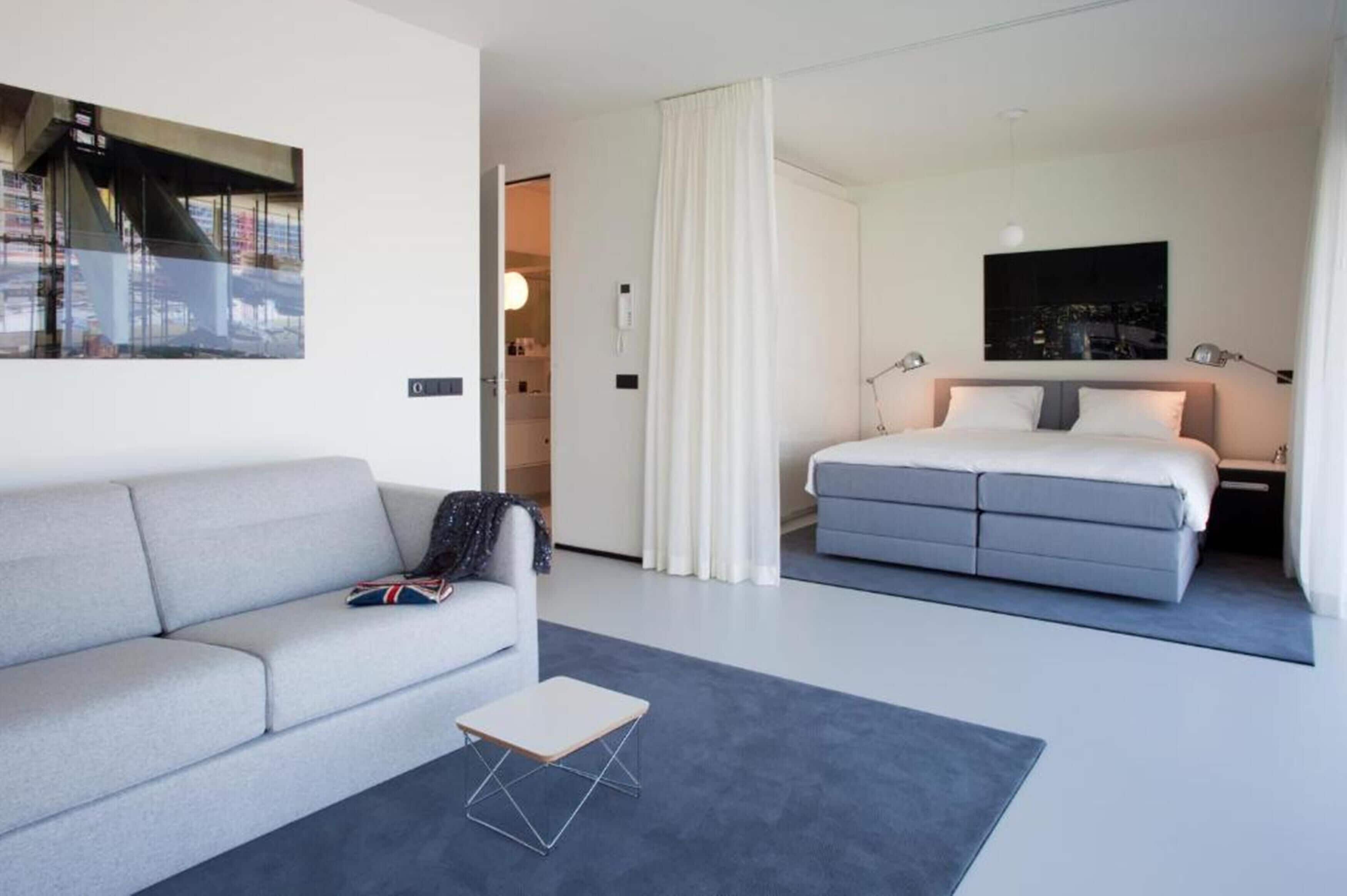 Ein modernes Hotelzimmer mit grauem Sofa, einem kleinen Tisch, einem Bett mit grauer Bettwäsche, Blick auf die Stadt und einem Gemälde an der Wand.
