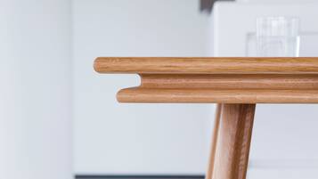 A wooden table with a simple design, positioned in a modern kitchen with white cabinetry and a glass cabinet in the background.