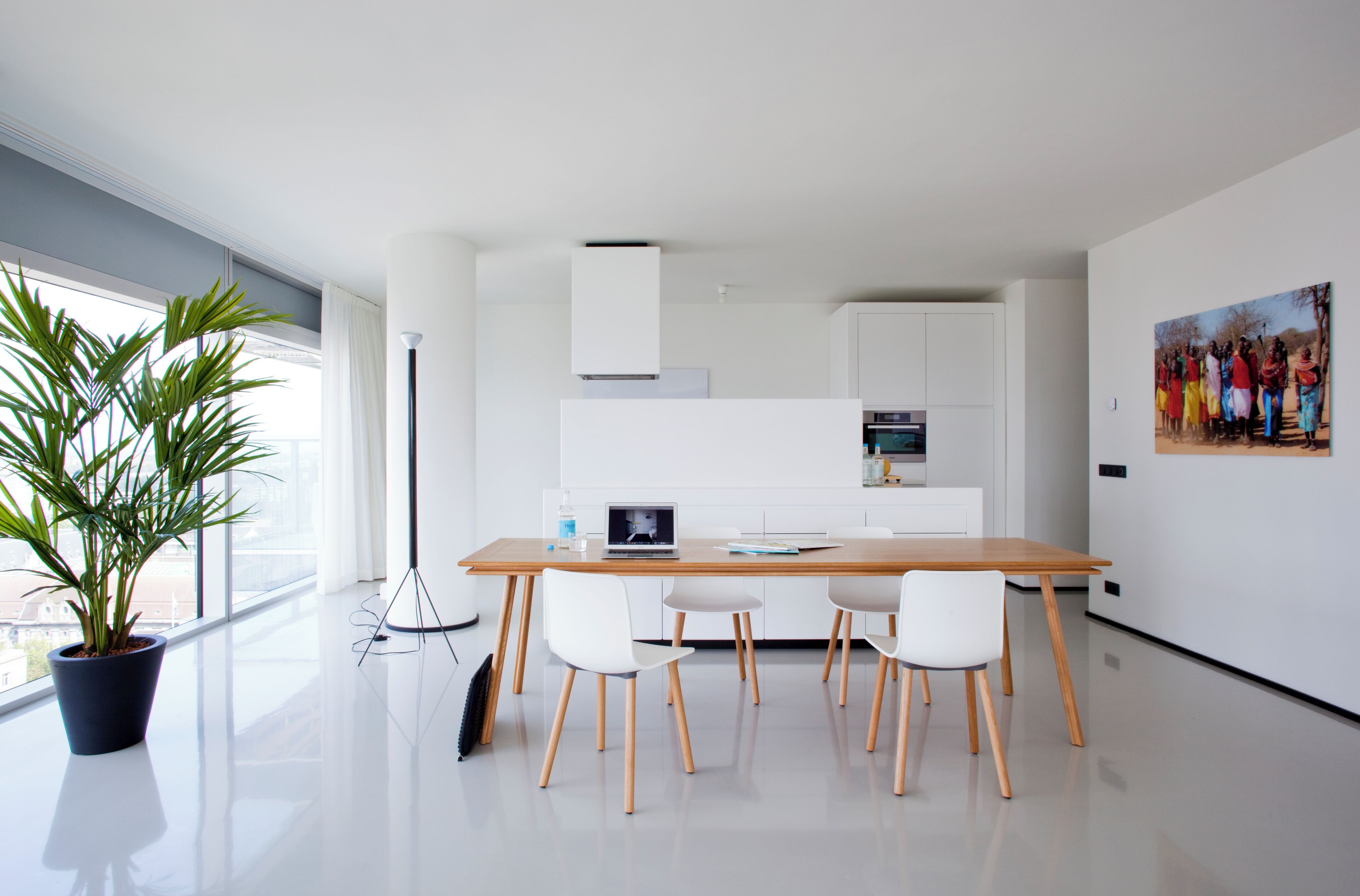 A modern kitchen with white cabinetry, a wooden dining table with four chairs, a laptop on the table, and a potted plant by the window.