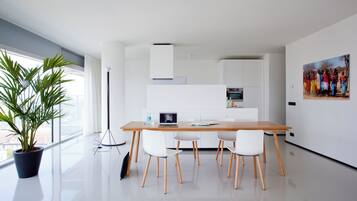 A modern kitchen with white cabinetry, a wooden dining table with four chairs, a laptop on the table, and a potted plant by the window.