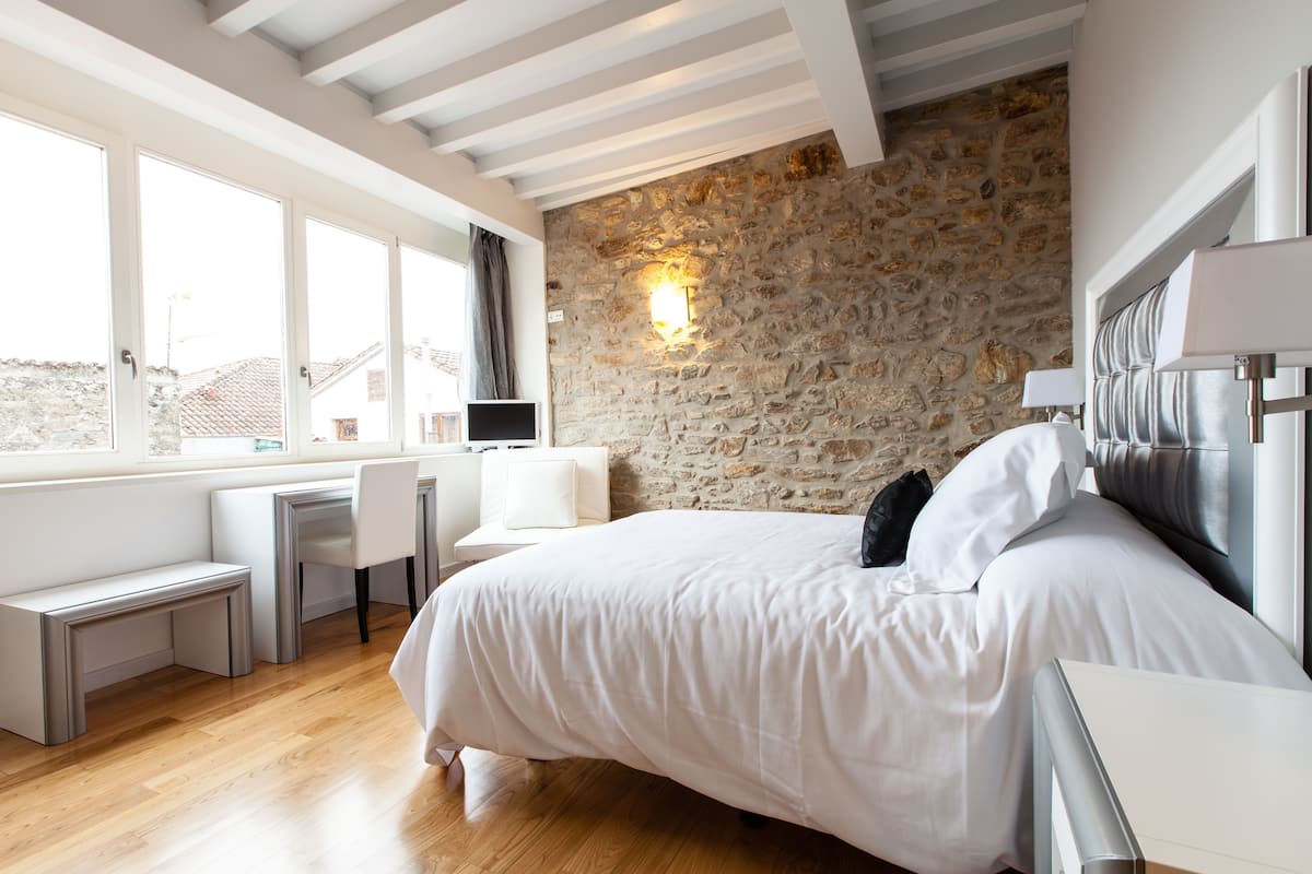 Een slaapkamer met een stenen muur, een bed met wit beddengoed, een bureau met stoel en een raam met gordijnen.