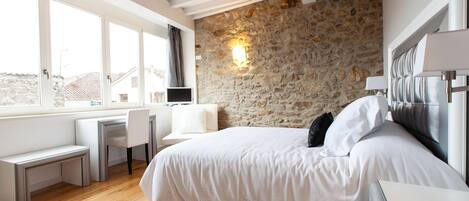 A bedroom with a stone wall, a bed with white bedding, a desk with a chair, and a window with curtains.