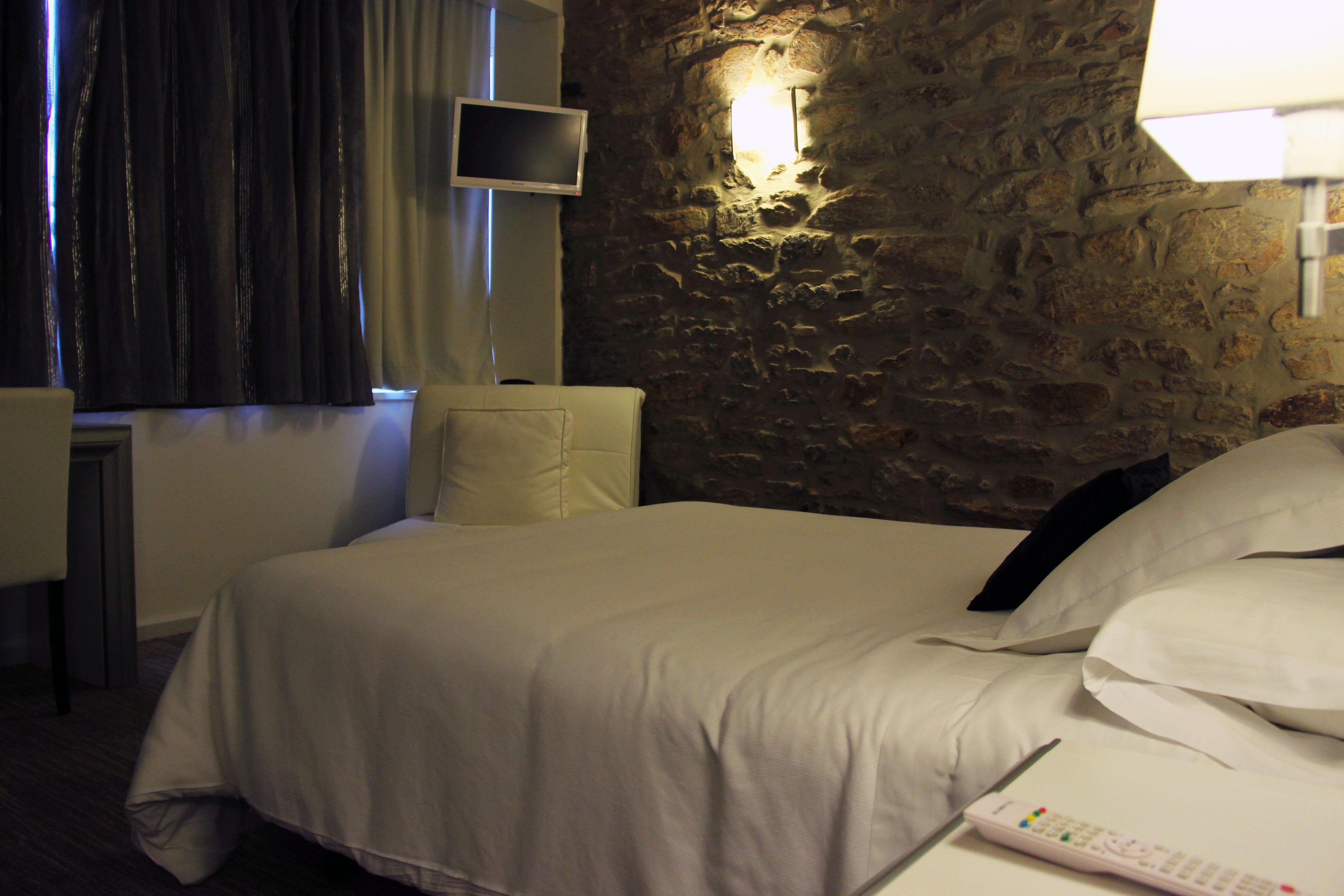 A hotel room with a stone wall, a bed with white bedding, a remote control on the bed, and a television mounted on the wall.
