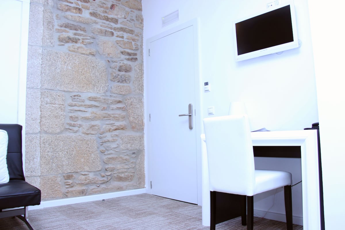 Een kamer met een stenen muur, een witte deur, een wit bureau met stoel en een wandgemonteerde televisie.