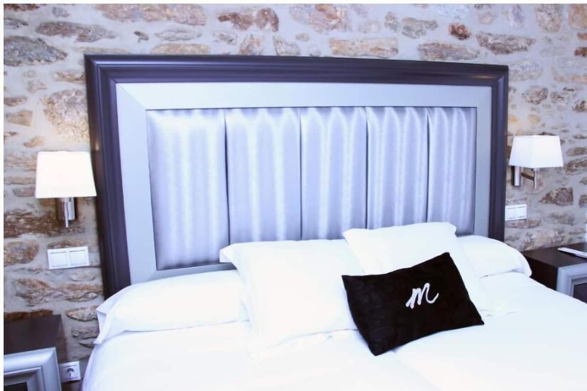 Een net opgemaakt bed met witte lakens en een zwart kussen met een wit 'M' logo, tegen een stenen muur met twee nachtlampjes.