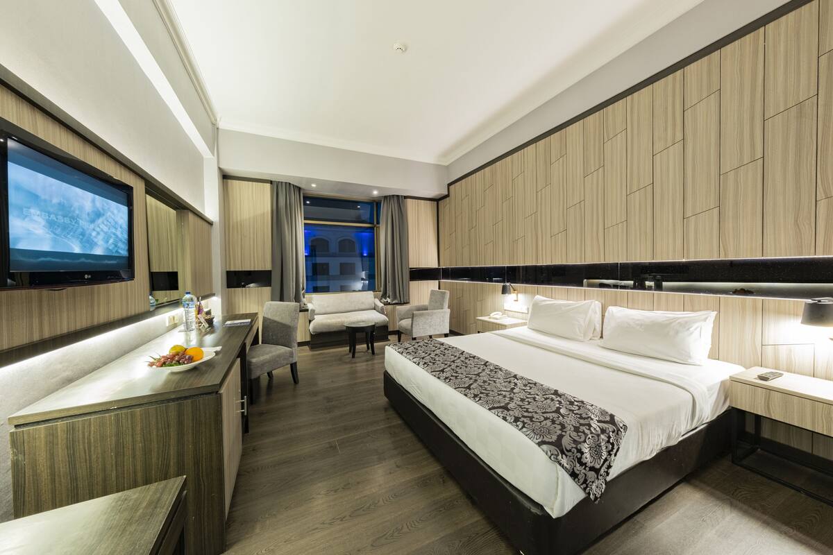 A modern hotel room with a large bed, a desk with a flat-screen TV, a sofa, and a view of the city through the window.