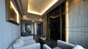 A modern hotel room with grey upholstered sofas, a wooden coffee table, and a flat-screen TV mounted on the wall.