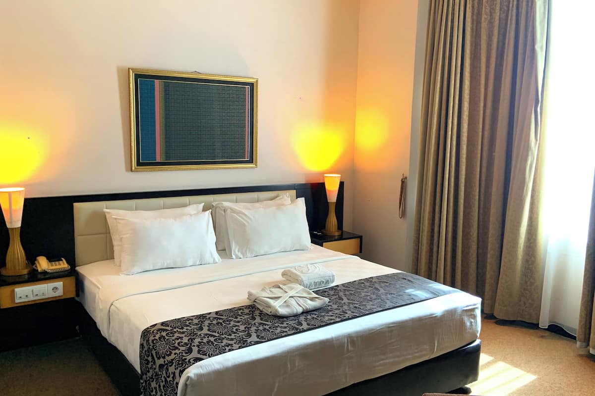 A hotel room with a bed, two bedside lamps, a nightstand with a phone, a framed picture on the wall, and a window with curtains.