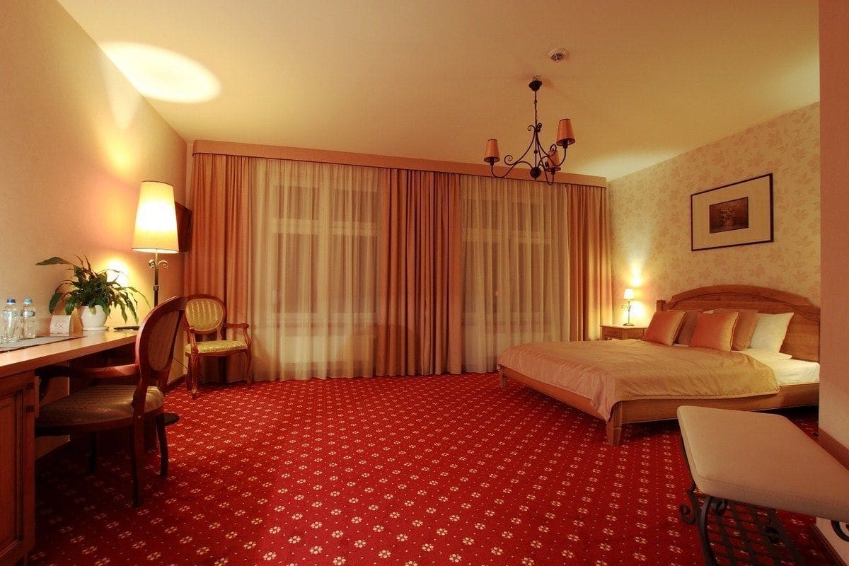 A hotel room with a bed, a desk, a chair, a lamp, and a chandelier.