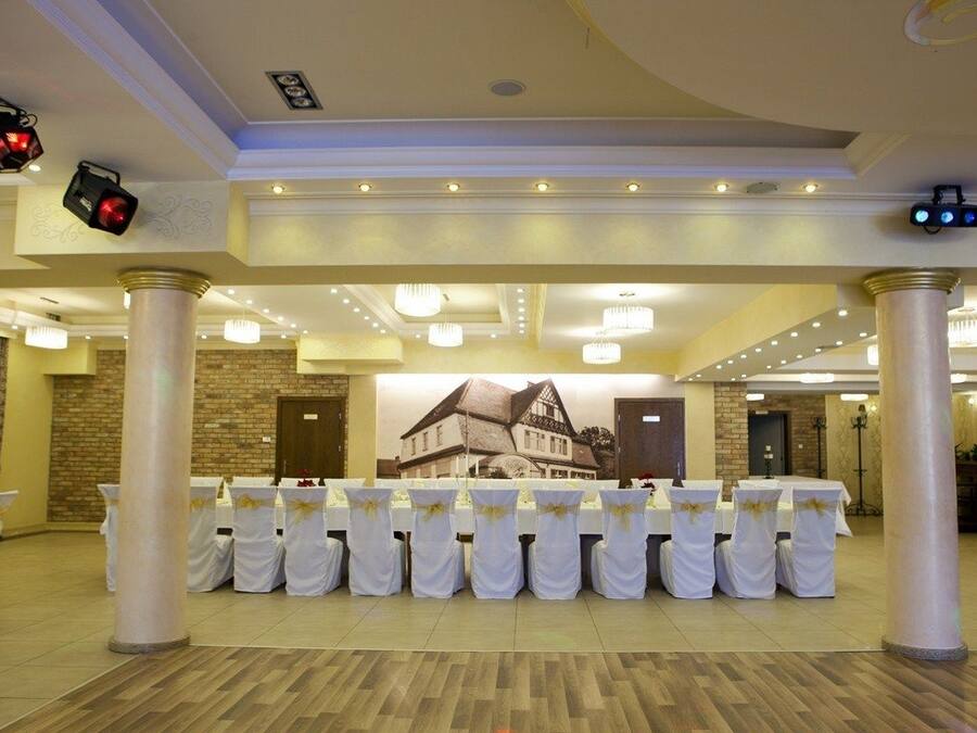 Un salón de banquetes con mesas y sillas blancas, un mural grande en la pared de fondo y una araña.