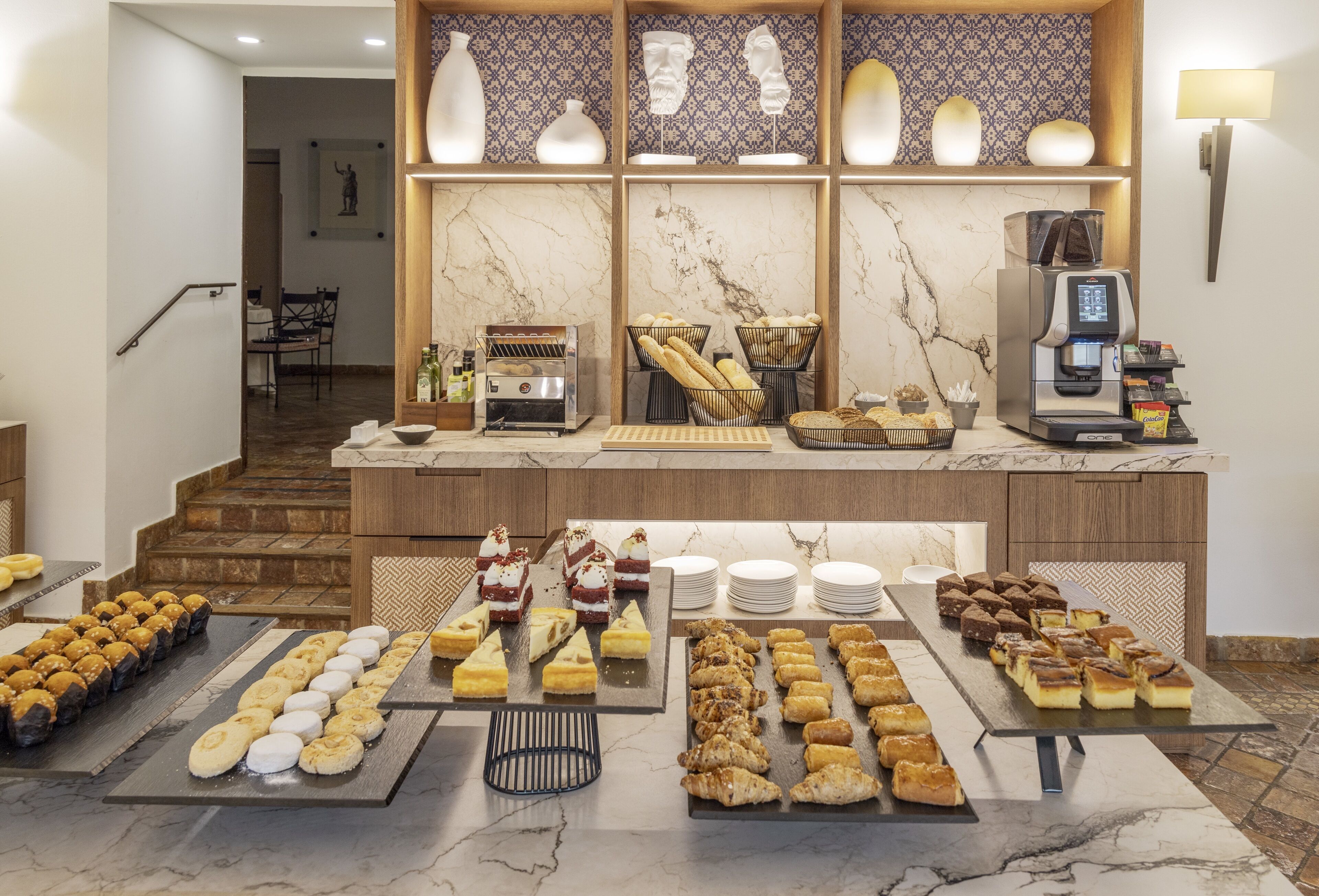 A buffet setup with various pastries and baked goods, a coffee machine, and a seating area with chairs and a table.