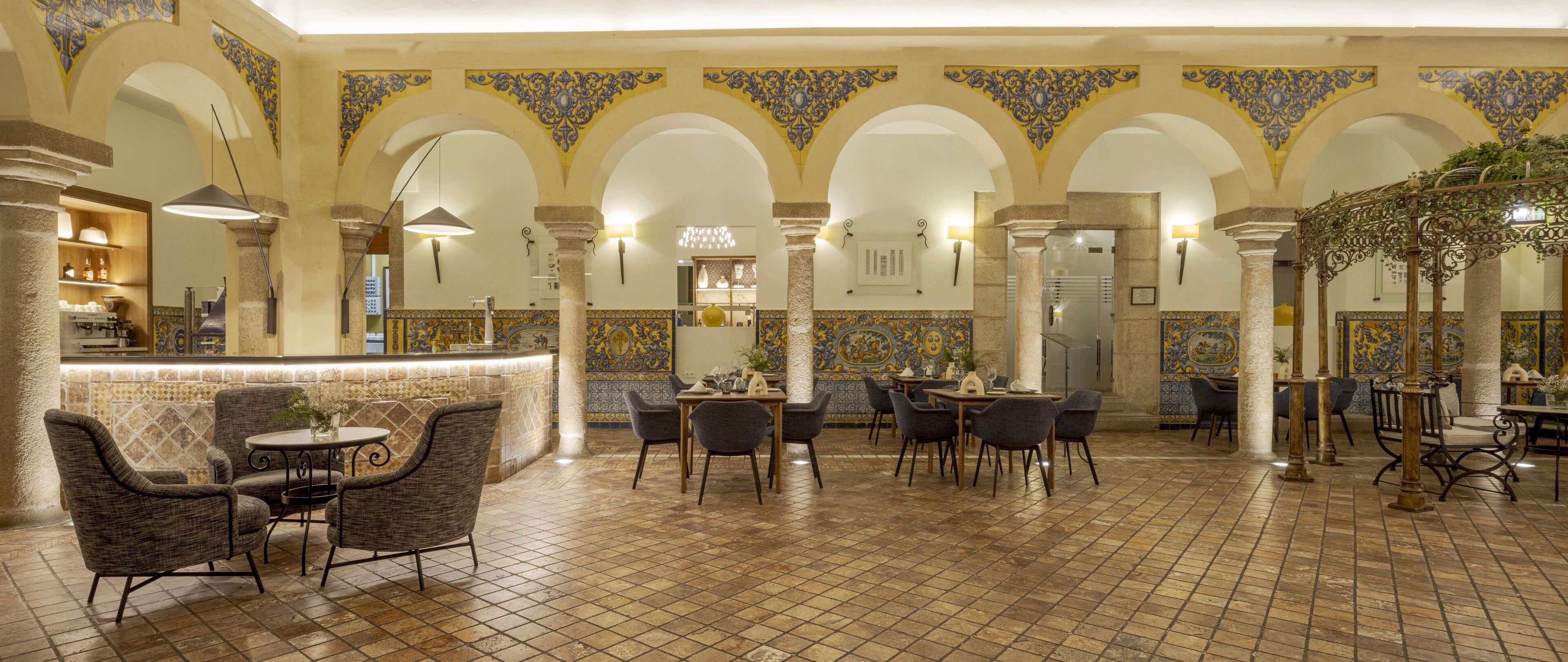 A spacious indoor area with a high ceiling, featuring a central seating area with round tables and chairs, surrounded by arches with decorative tile work.